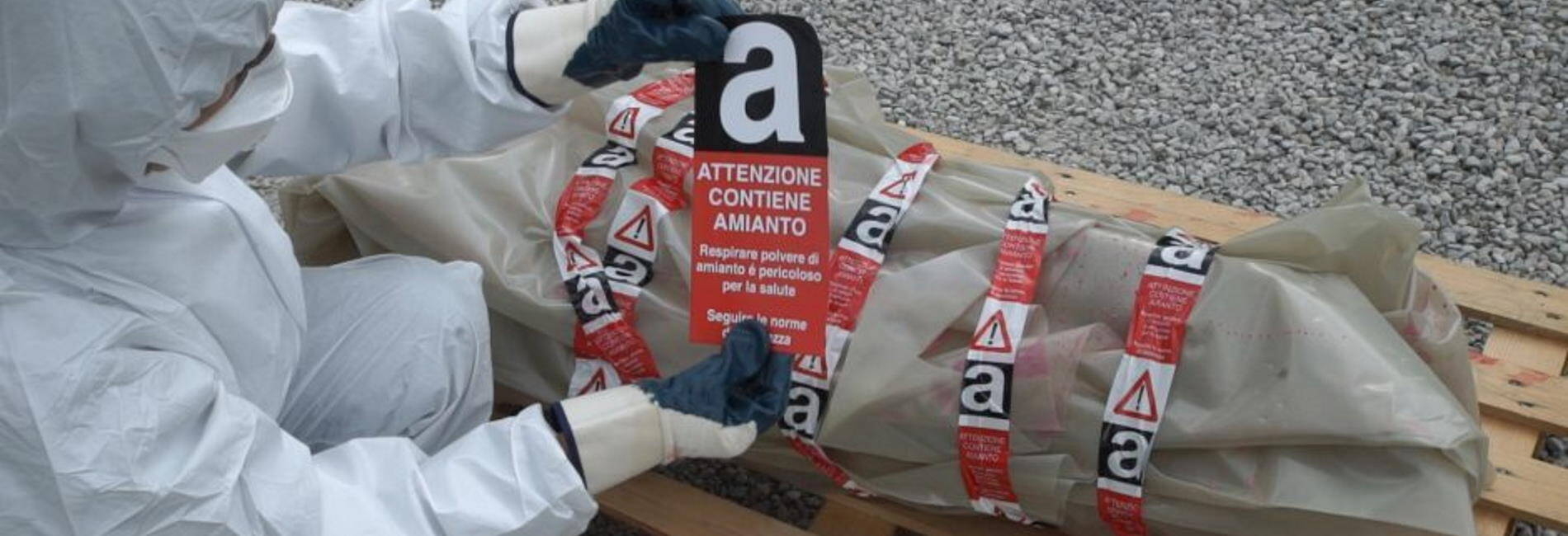 Persona con tuta di protezione e guanti che sta attaccando un adesivo "Attenzione contiene amianto" su del materiale impacchettato 