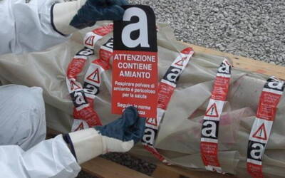 Persona con tuta di protezione e guanti che sta attaccando un adesivo "Attenzione contiene amianto" su del materiale impacchettato 